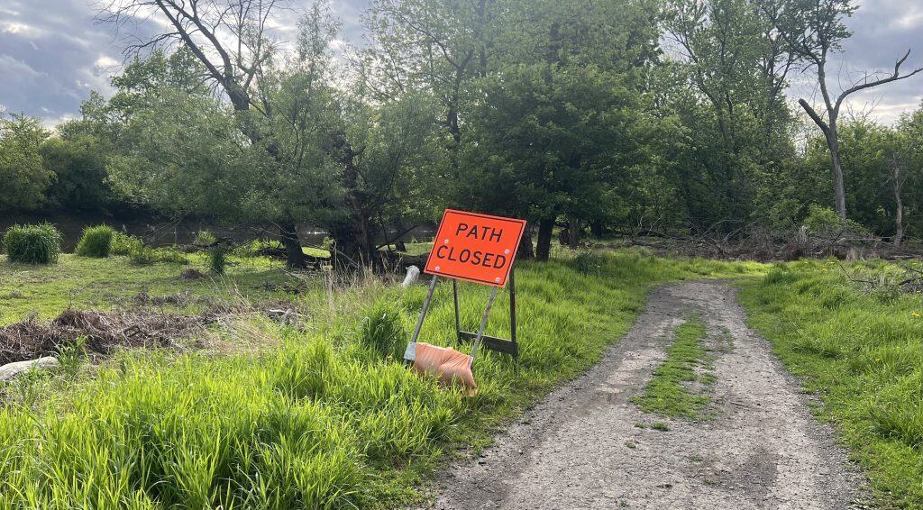 Looking at an orange Path Closed sign in grass to the left of a muddy trail of crumbled asphalt, dirt and grass, and with a riverfront, the river and trees in the background.