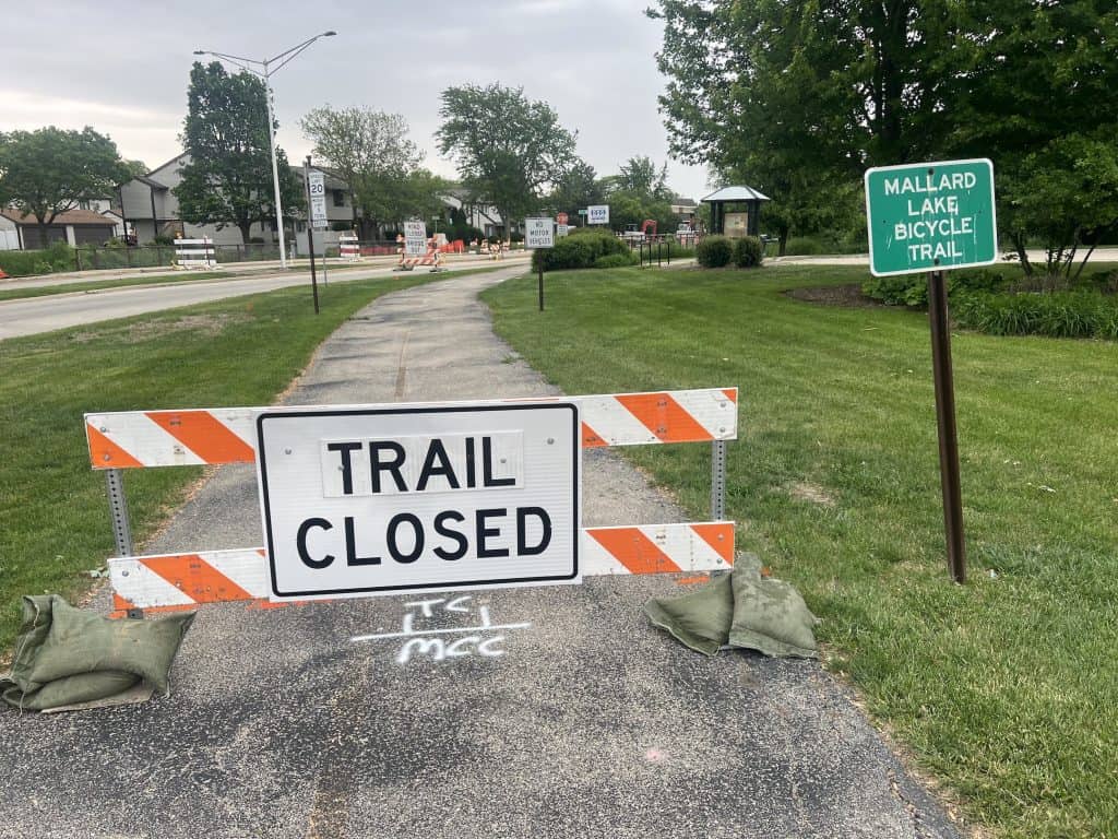 A construction barrier with a Trail Closed sign blocks a trail with a sign to the right labeling it the Mallard Lake Bicycle Trail.
