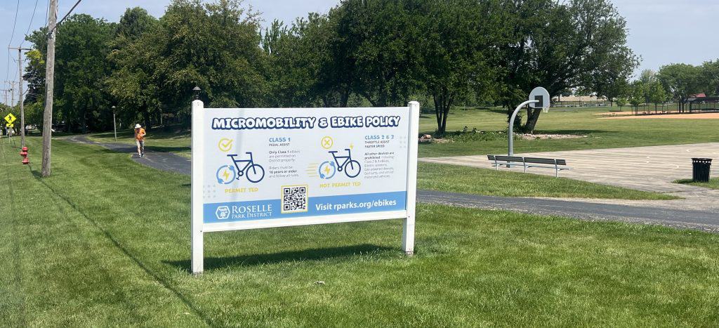 A big sign about e-bike policy stands on the lawn on the side of a suburban park.
