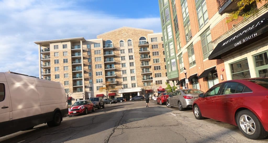 Looking down a street in the middle of a suburban downtown, with tall new buildings to the right and ahead, and cars parked around.