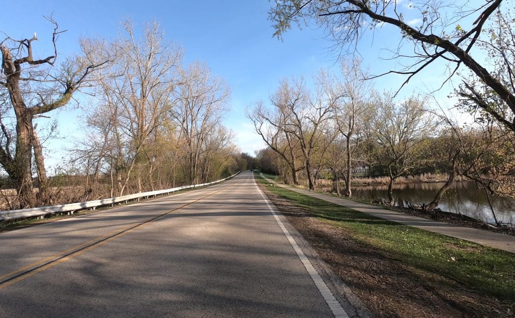 Looking down a two-lane road in a leafless tree-lined area, with a parkway and a sidewalk on the right.