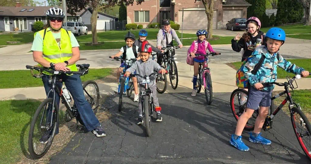 An adult and several children on bikes, wearing helmets, post in a suburban neighborhood on a paved path.