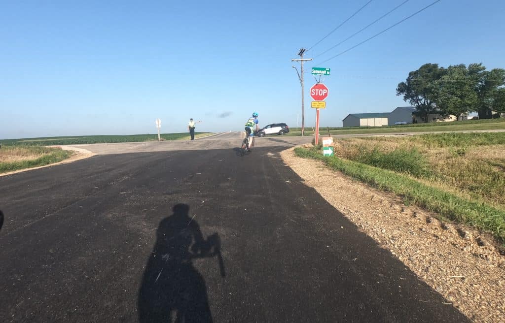 A shadow of a cyclist appears up front on a new asphalt road, with a sheriff's deputy pointing to the right at an intersection where another cyclist turns ahead of a stop sign, in wide open farmland.