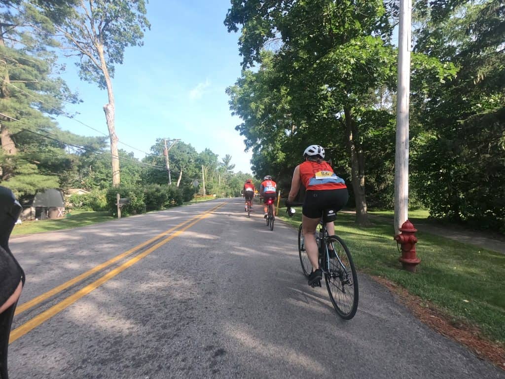 A few cyclists ride on a road through a tree-filled suburban neighborhood.