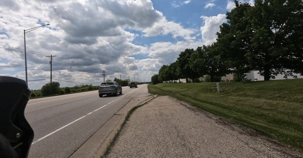 A path before us is about to end and blend into the shoulder of a wide road with lawn and short trees on the right side.