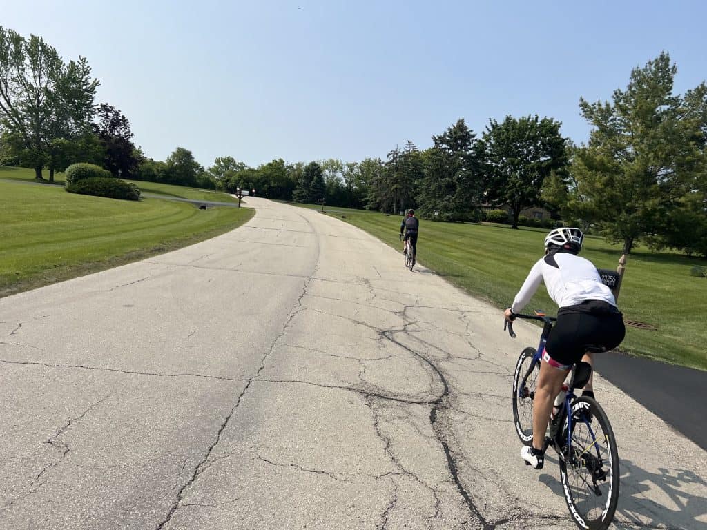 Two cyclists ride on a wide road through a spacious suburban neighorhood with lawn and short trees.