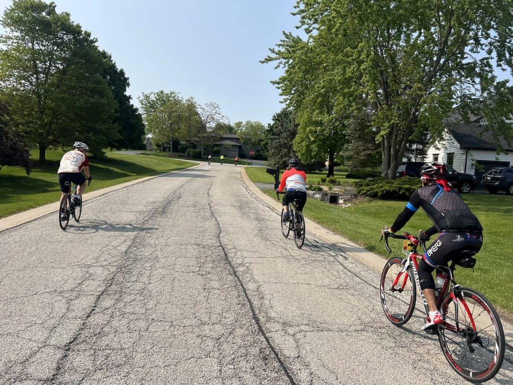 A half dozen spread-out cyclists ride through a spacious suburban neighborhood with trees.
