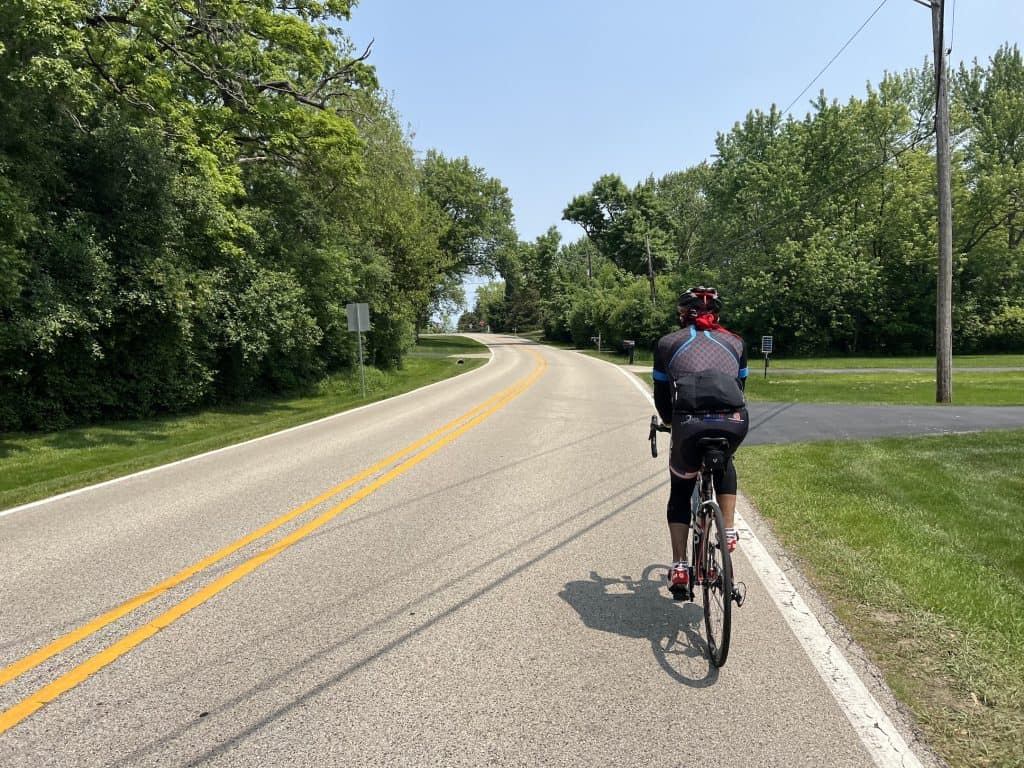 A cyclist rides on a paved two-lane road through a tree-lined rural area.