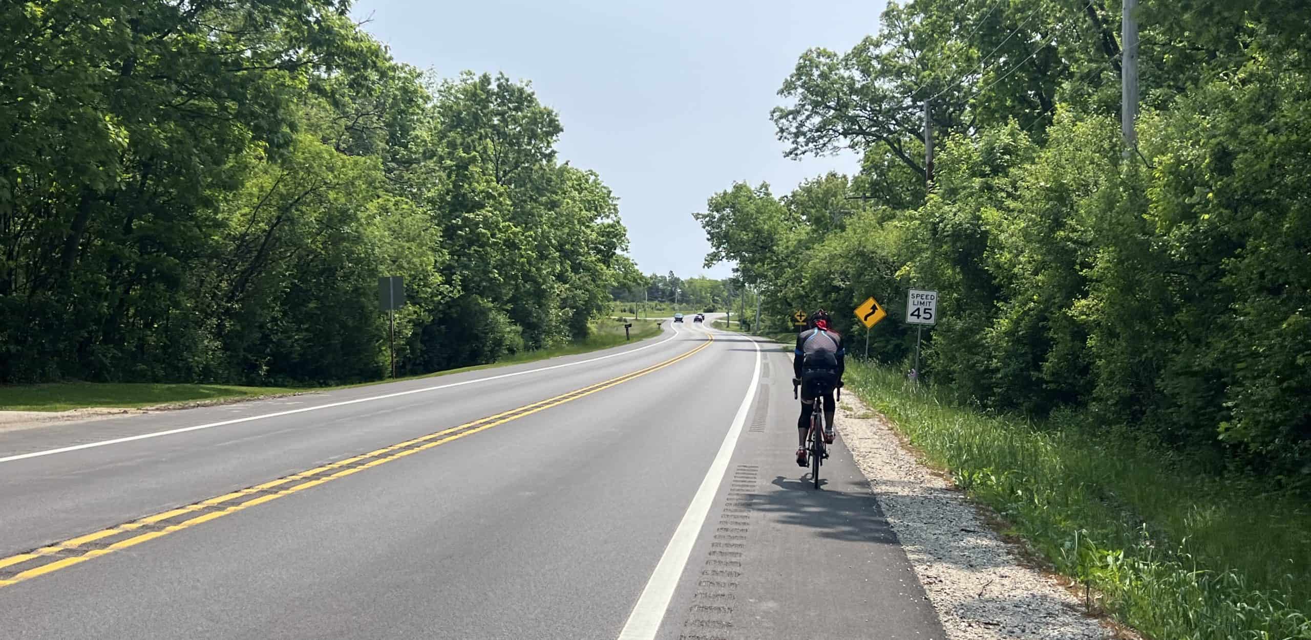 Looking down a paved two-lane road with a cyclist up ahead on the shoulder, trees lining both sides.