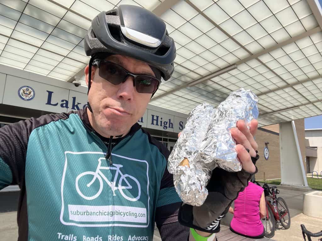 A cyclist poses with two hot dogs wrapped in aluminum foil in front of a high school entrance.
