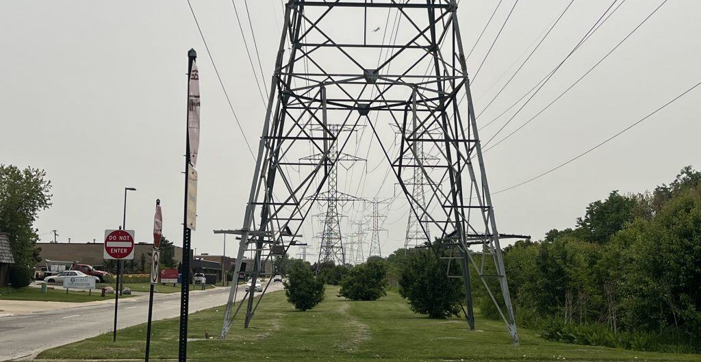 Looking down a series of electric towers with power lines over a wide median with grass and shrubs, with a street crossing up front and two-lane streets on the left and plane in the hazy sky seen through the closest tower.