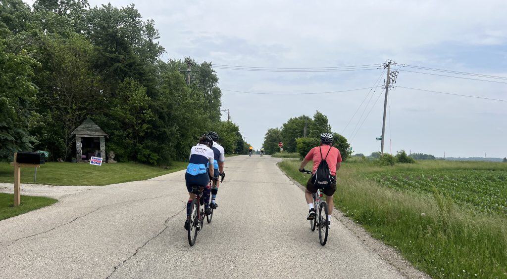 Bicyclists ride an open unstriped road by full trees on the left and prairie with power lines on the right.