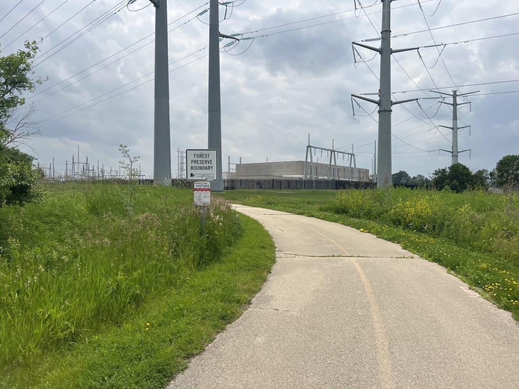 A striped, older paved trail curves left through grassland with power lines and towers in the background and a Forest Preserve Property sign.