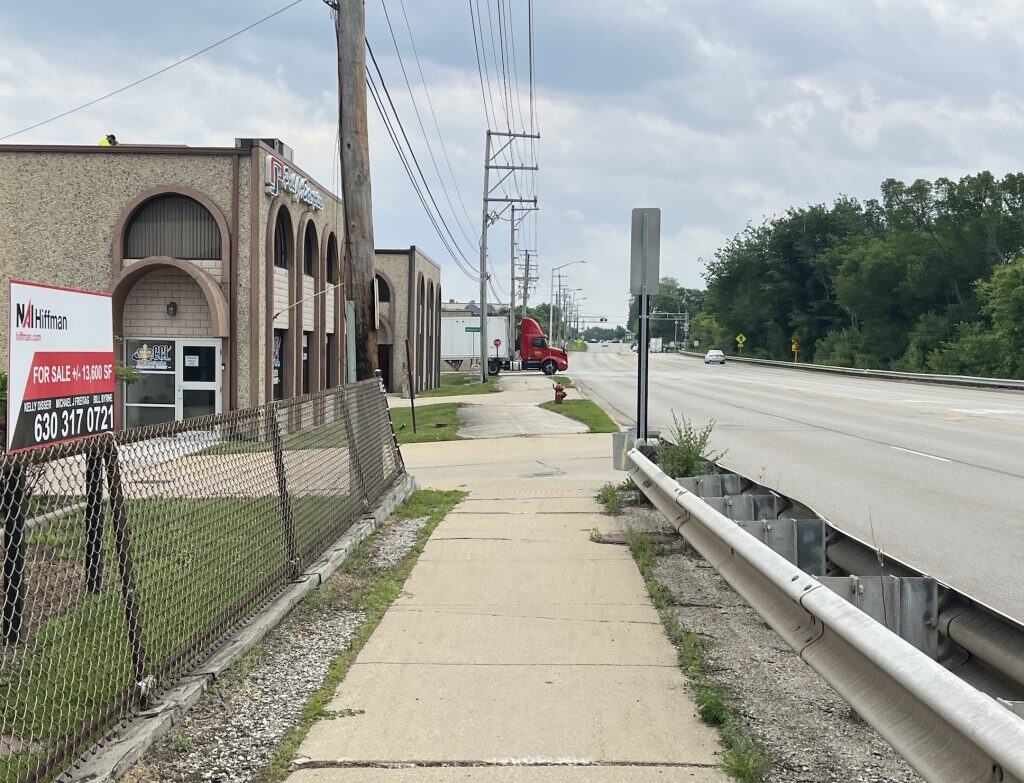 A sidewalk ahead crosses a driveway, then becomes a striped bike path before another driveway and finally a street up ahead with a semi truck turning out of it, with industrial buildings on the left and a four-lane road on the right.