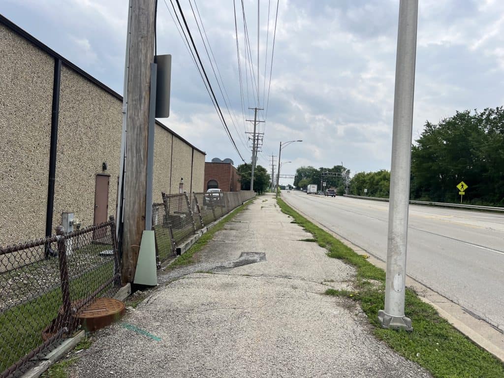 Looking down a bumpy paved path past an industrial building on the left and a four-lane road on the right, under power lines.