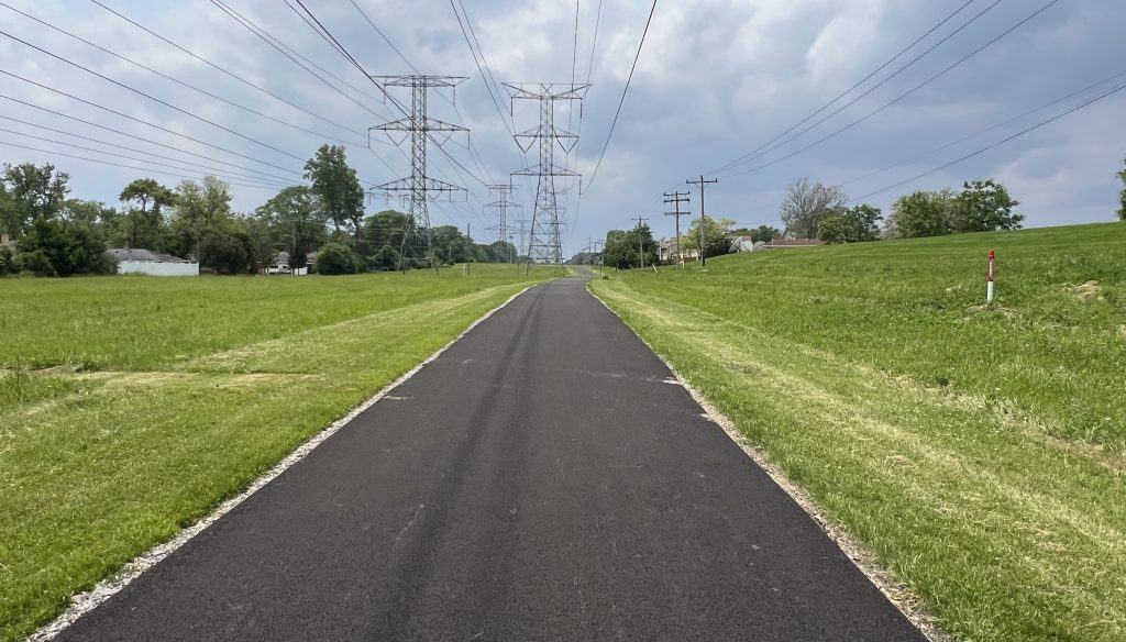 Looking down a freshly made black asphalt path through grass and under power lines.