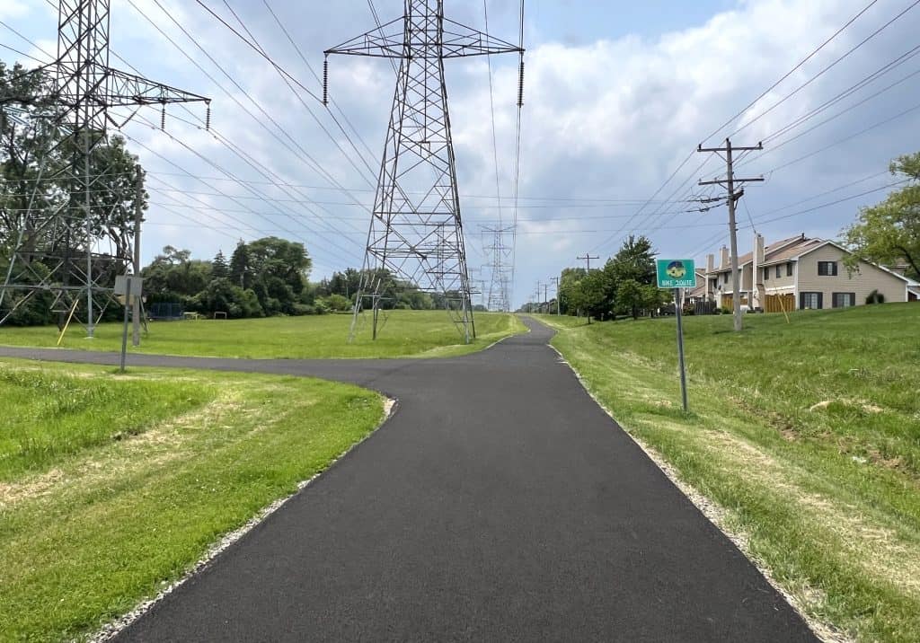 Looking down a new black asphalt path through a greenway under power lines, with a segment curving left and a Bike Route sign on the right.