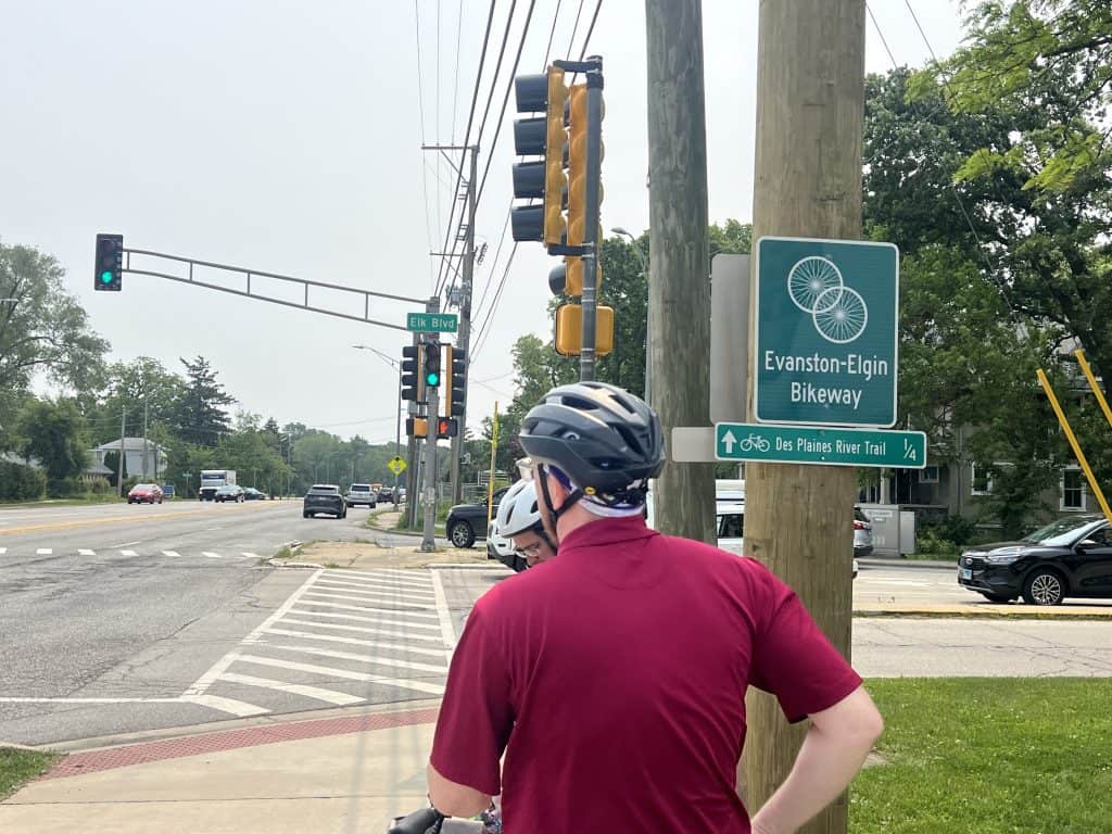 Bicyclists stop at a busy suburban intersection where there are traffic lights and crosswalks on pavement.