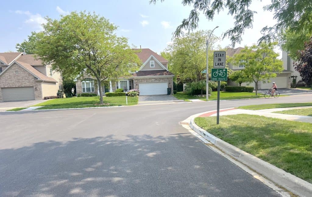 A bicyclist in the far right background prepares to turn on newly paved curving street through a suburban neighborhood.