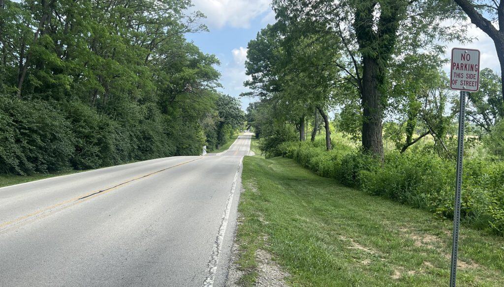 Looking down a paved road lined with trees and a prairie in the background on the right.