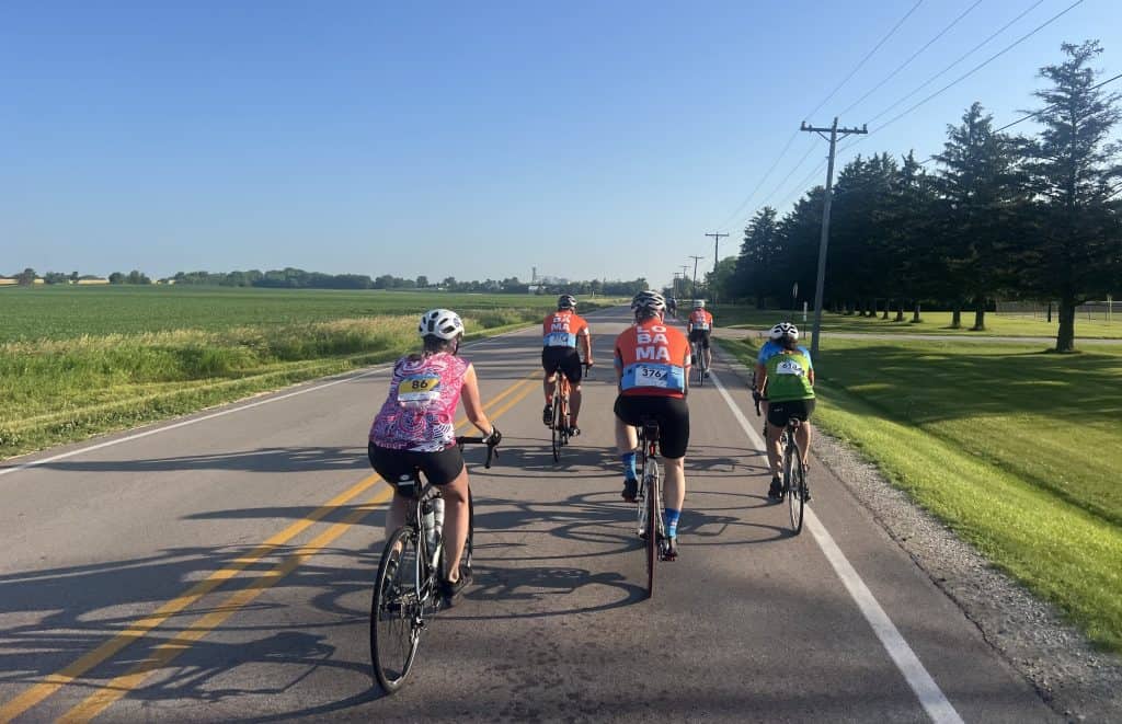 Several cyclists ride down a two lane open road with green farmland on the left, and power lines and trees on the right.