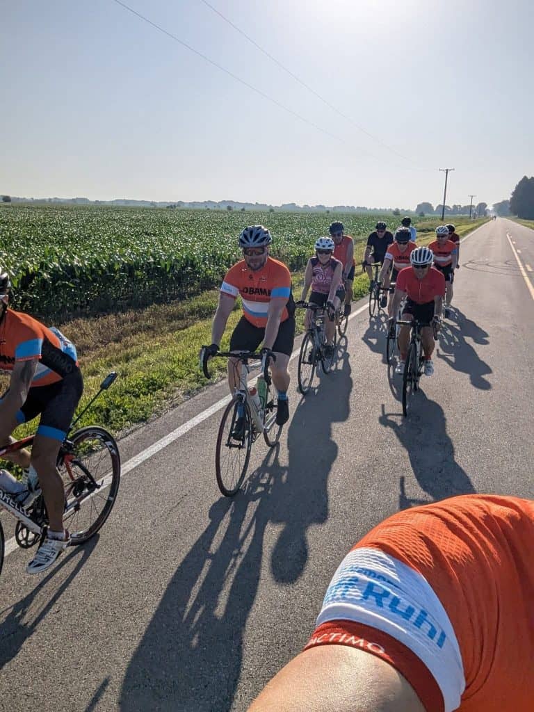 Several cyclists ride toward us on an open road through a field of growing green corn stalks under a bright sun.