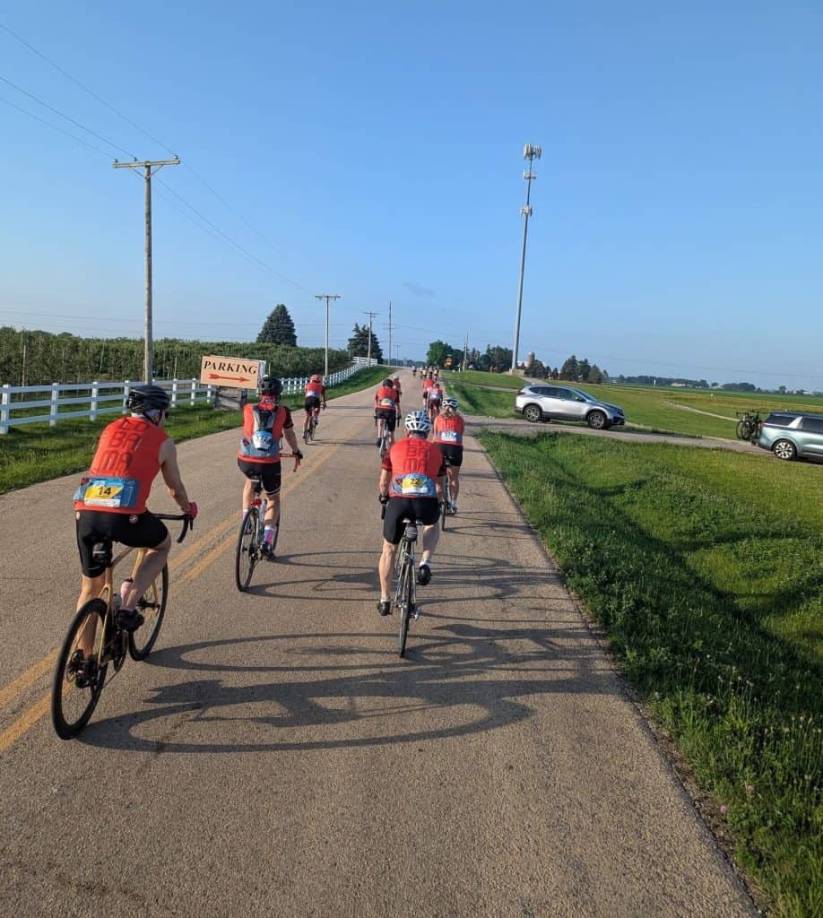 A group of cyclists in orange jerseys head down a road between farms, with cars parked on the right.