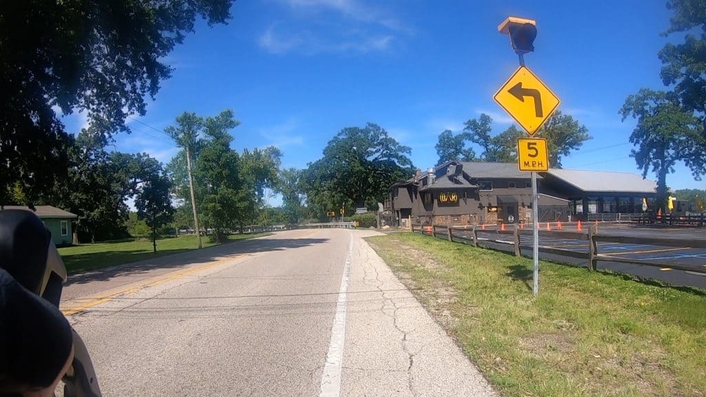 Looking down a two-lane road about to curve left, with a 5mph left-turn arrow sign up front, a house-like structure on the right, trees on the sides and a river in the background.