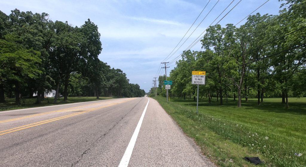 Looking down the shoulder of a two-lane road through an exurban area with short trees on the sides, and a Lake Barrington village sign and a couple of other signs.