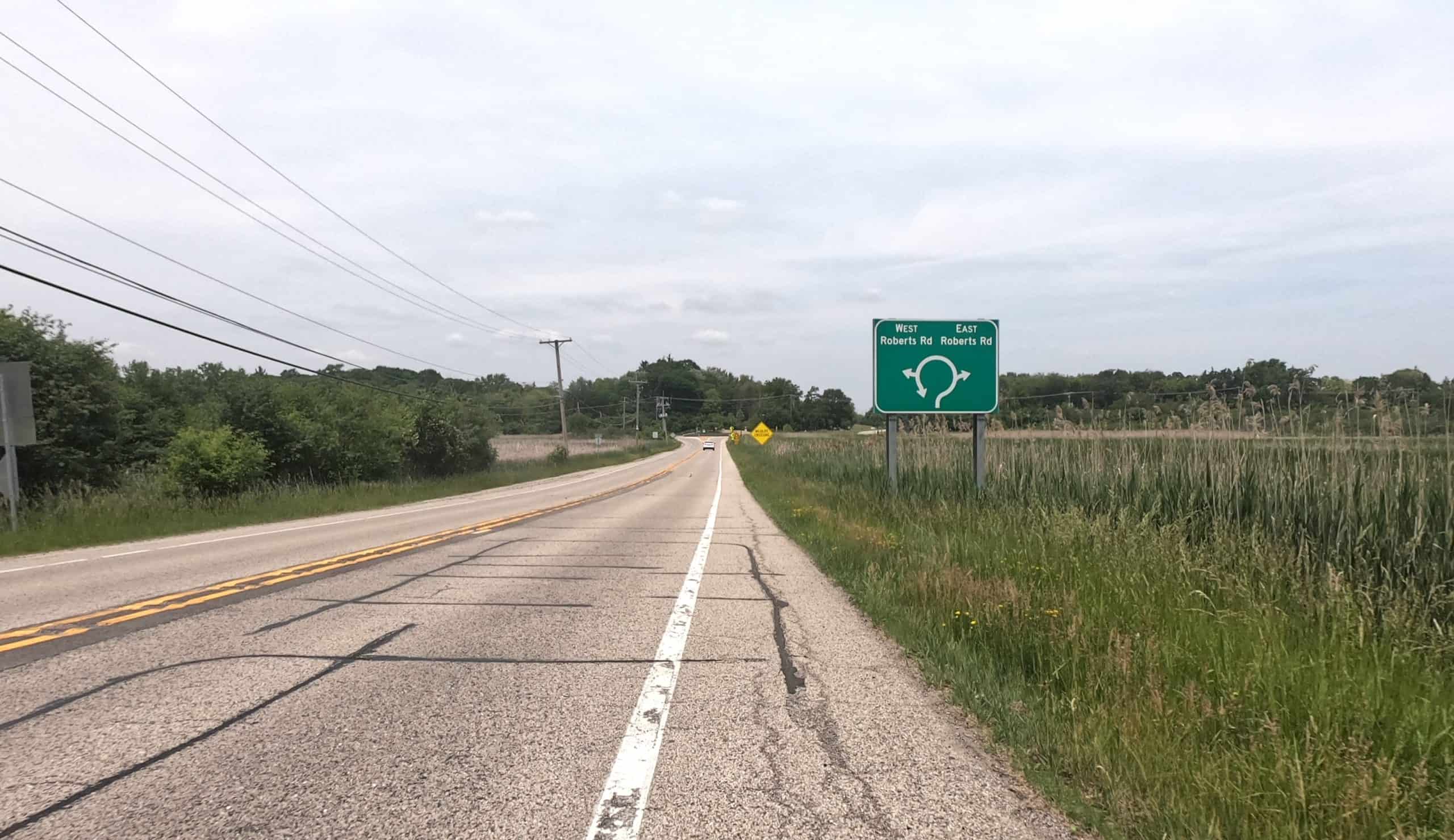 Looking down the shoulder of a two-lane road through prairie, trees in the background and a sign indicating a coming roundabout.