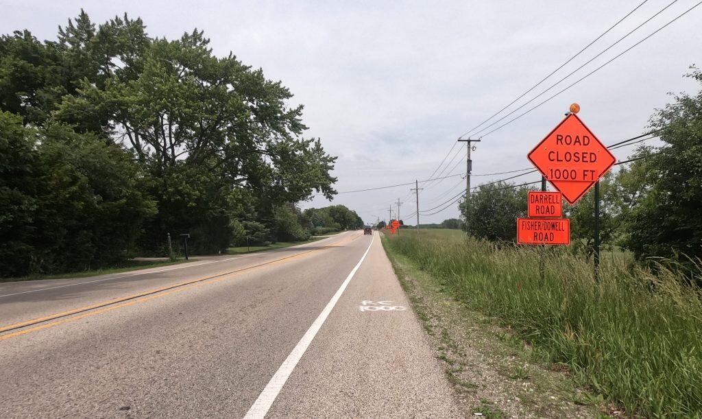 Looking down the shoulder of a two-lane road with tall grass and prairie on the right and trees on the left, with orange Road Closed signs up front.