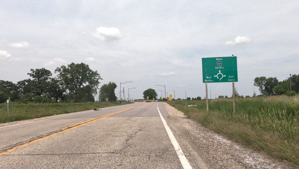 Looking down a two-lane road through prairie with a few trees in the background left, and a sign up front indicating a roundabout coming up.