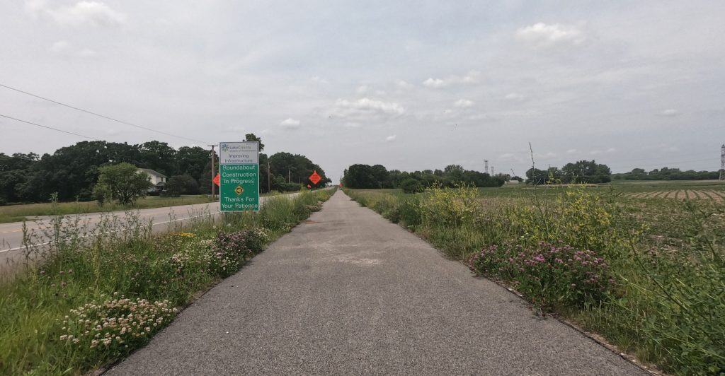 Looking down a paved path alongside an open road through prairie with colorful prairie flowers, and a sign indicates a roundabout project.