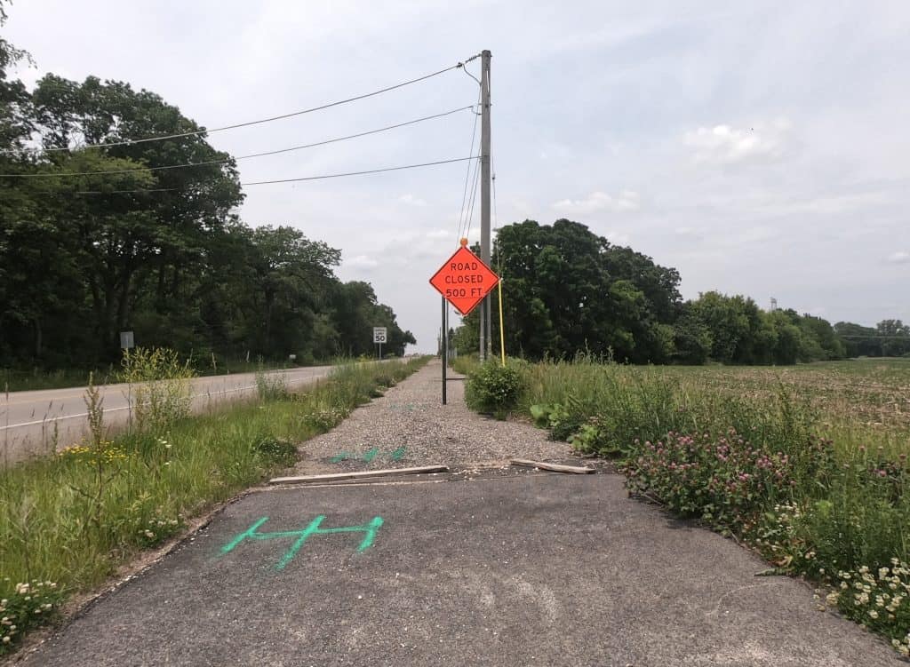 A paved trail through prairie and alongside a road ends with a gravel trail and Road Closed sign behind it, trees in the background.