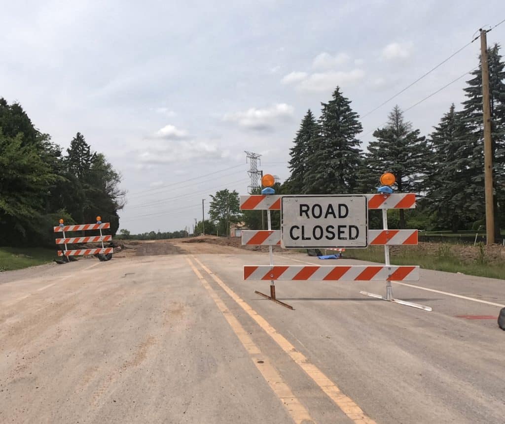A Road Closed construction barrier stands at the end of a two-paved road ahead of dirt being torn up behind it.