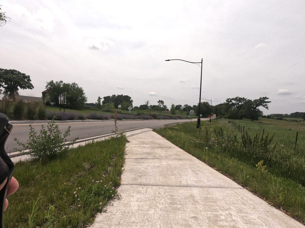 A concrete path ends at an asphalt road that continues slightly to the right ahead, under a streetlight through open prairie and scattered trees.