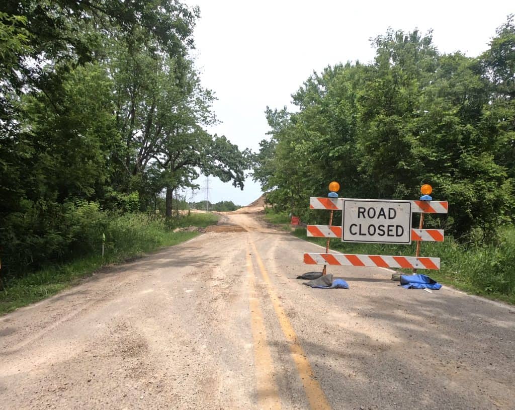 A Road Closed sign on a construction barrier stands on a dirty paved road with trees and a dirt road in the background.