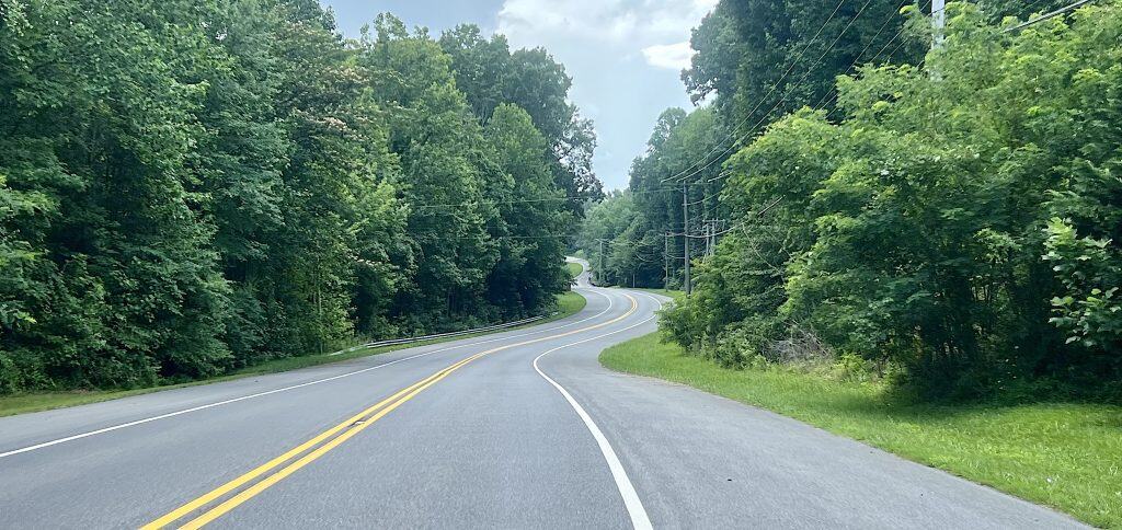 Looking down a curvy two-lane paved road with a shoulder through a forested area.