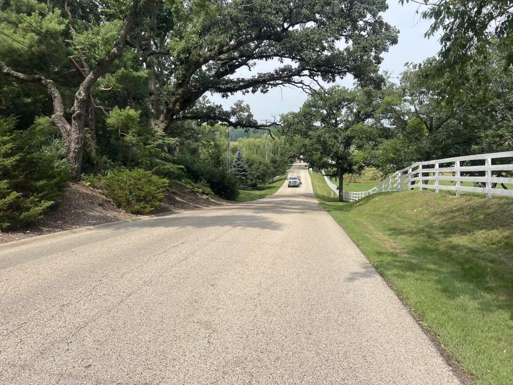 A road goes down a hill with tall trees overhanging on the left, a white fence on the right and a truck up coming up ahead.