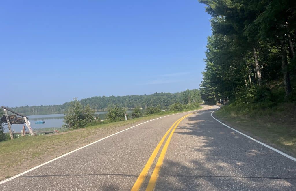 A two-lane road curves to the right past tall pine trees on the right and a pond with trees in the background on the left.