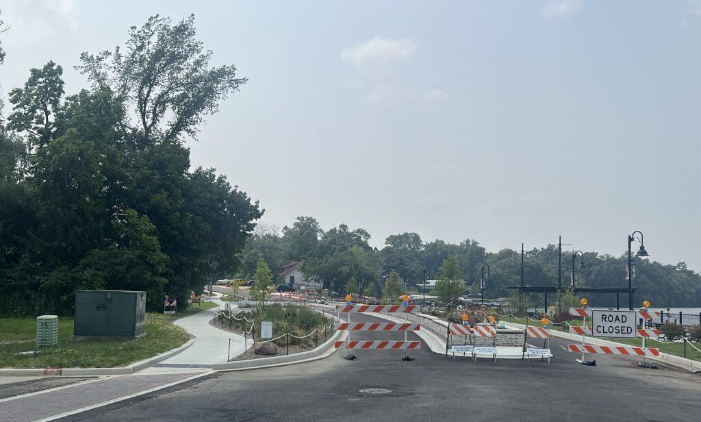 Looking at a construction scene with construction barriers up front, new asphalt on the road and new sidewalks on the sides, with a new median with flowers in the middle, trees and a lake in the background.