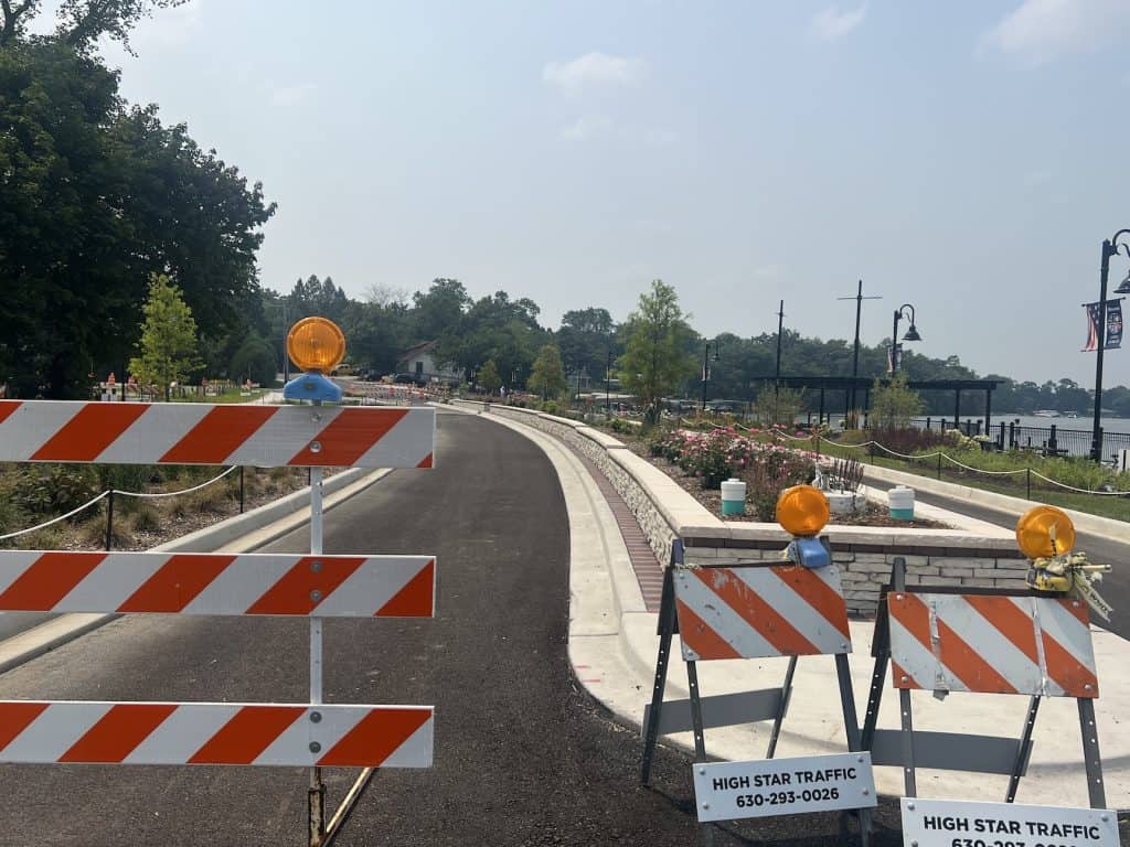 Looking past construction barriers at a newly paved, narrow road alongside a median with flowers.