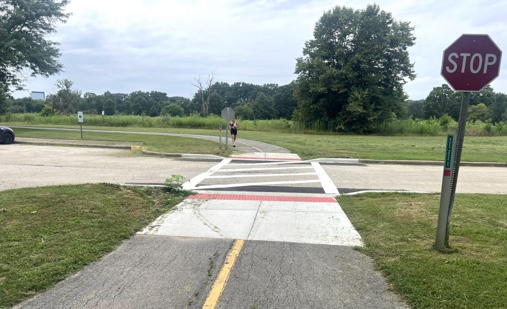 Road crossing at a suburban bike trail with a stop sign, runner, and green natural surroundings.