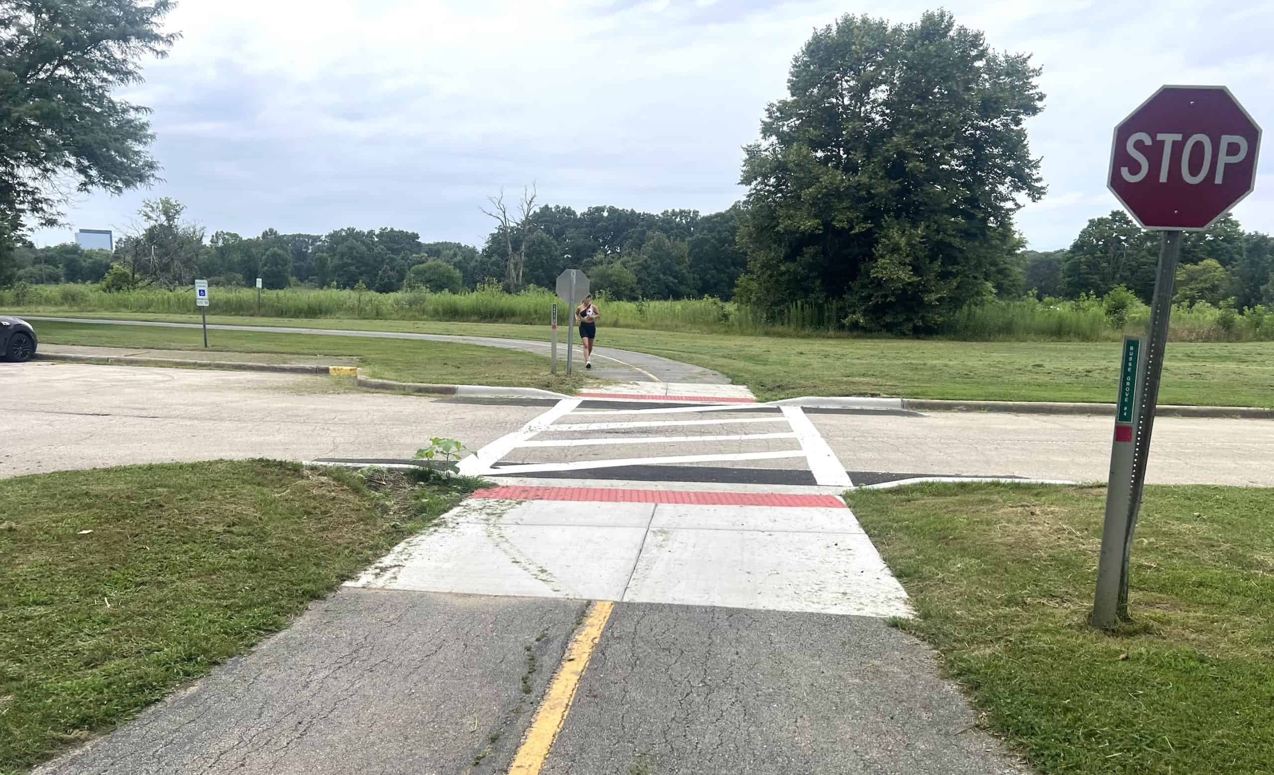 Road crossing at a suburban bike trail with a stop sign, runner, and green natural surroundings.