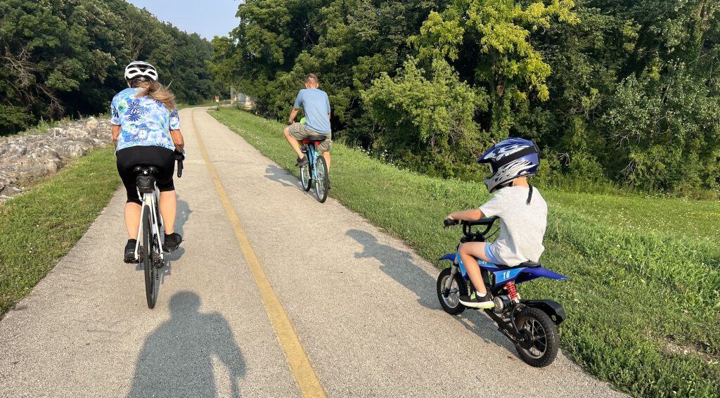 A few bicyclists ride on a forest preserve striped bike trail, with a boy on a small motorized mini-bike upfront on the right.