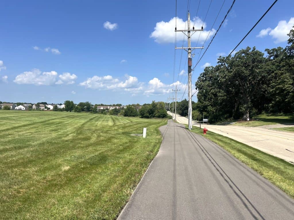 An asphalt bike path runs along a meadow on the left and a street on the right in a spacious suburb under a blue sky with a few white clouds.