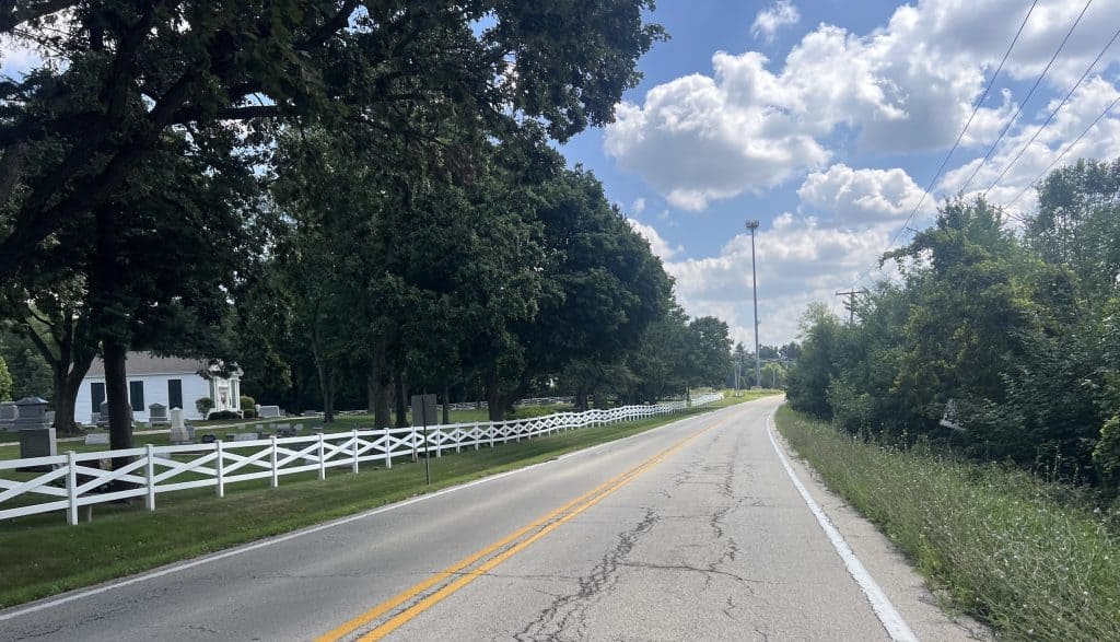 Looking down a 2-lane road curving slightly right past a white picket fence on the left with tall trees lining the left side, under a sunny partly cloudy sky.