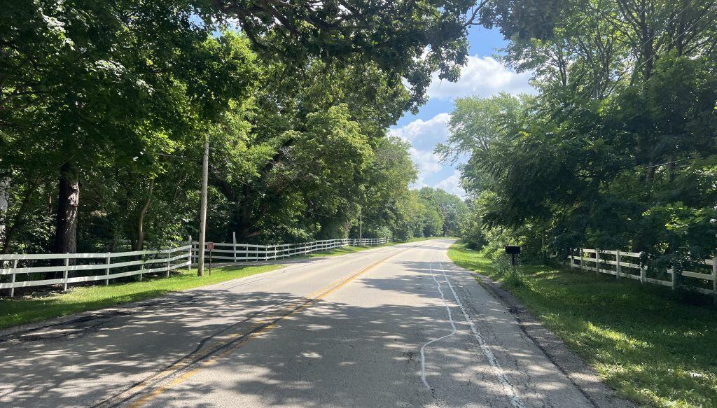 Looking down a 2-lane road curving to the right in the country on a sunny day, with a white picket fence on the left and tall trees lining the road.