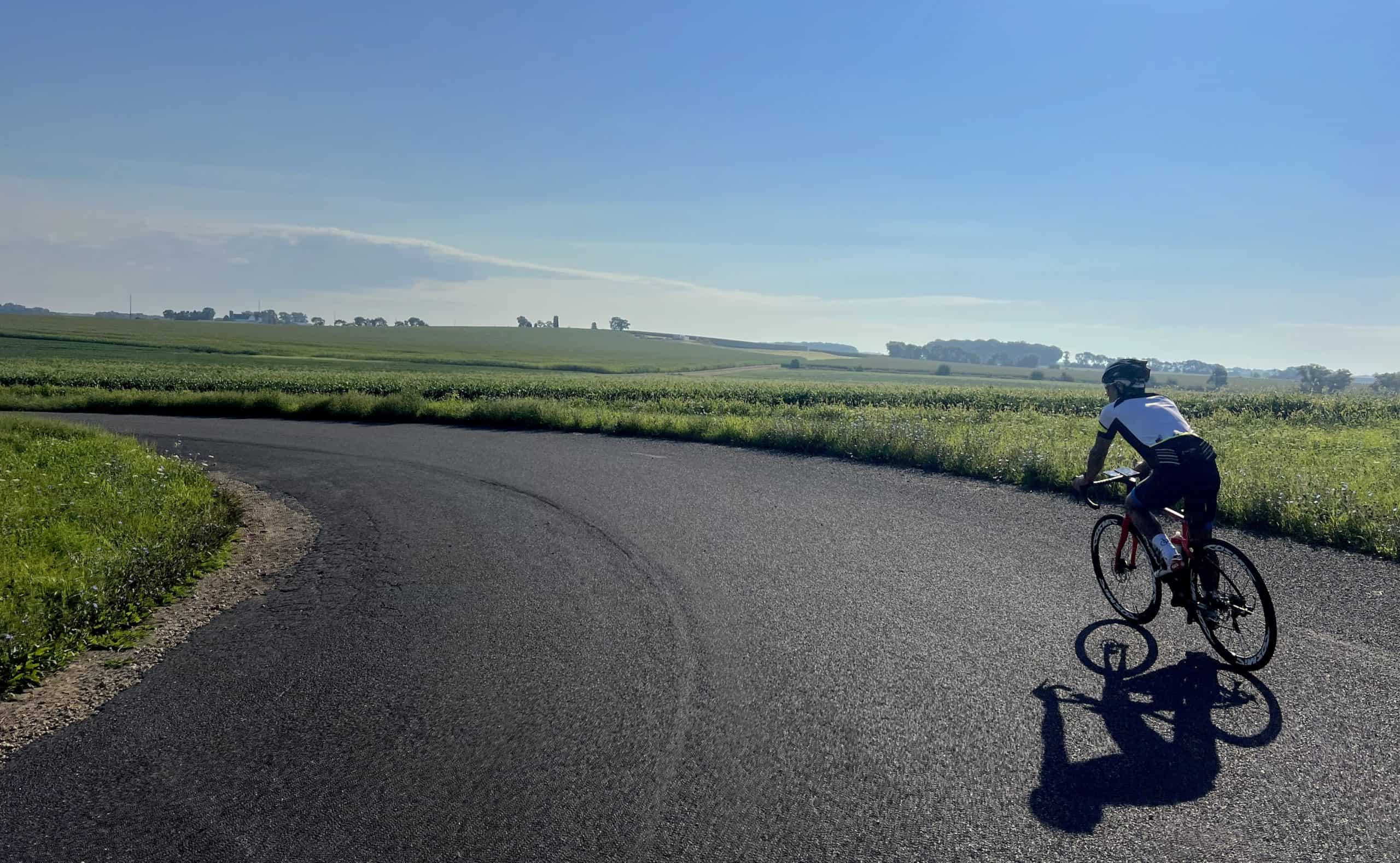 A cyclist and his long shadow turn on an asphalt road turning sharply left in front of slightly hilly farmland, under a blue sky.