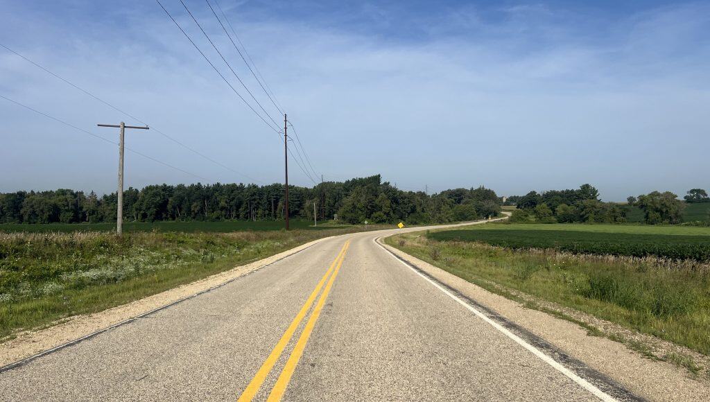 A two-lane road curves through farmland with power lines on the left side and trees far in the background under blue sky.